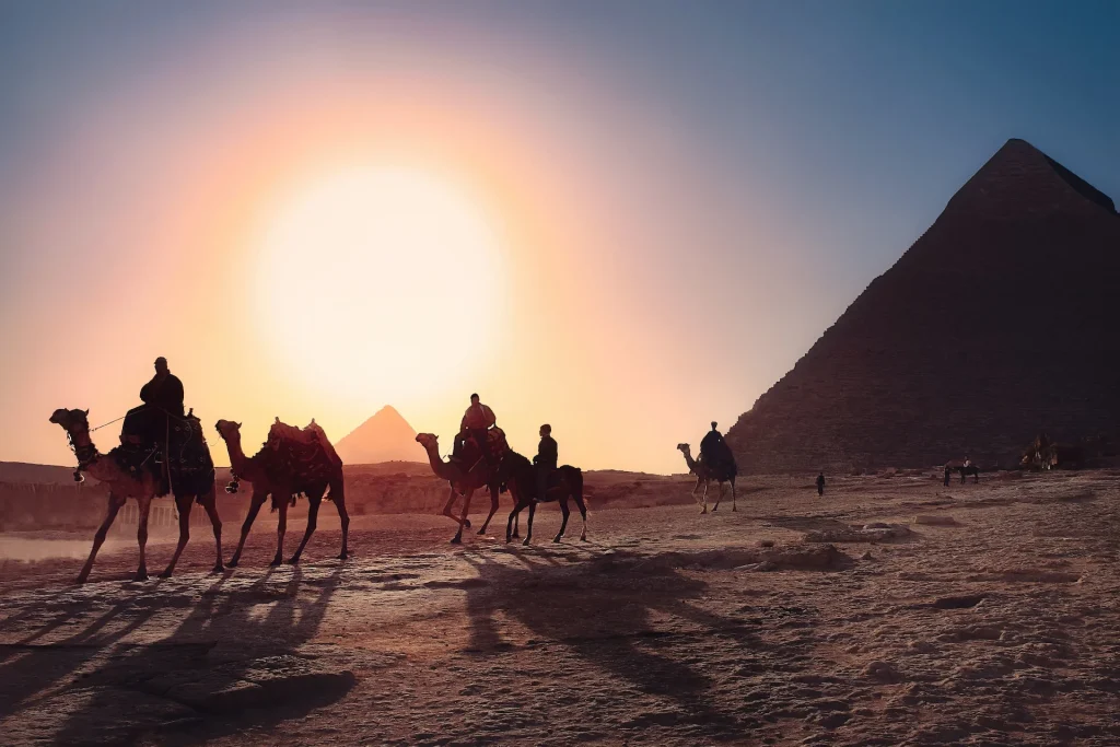 Cinco pessoas andando de camelo andando na areia ao lado da Pirâmide do Egito
